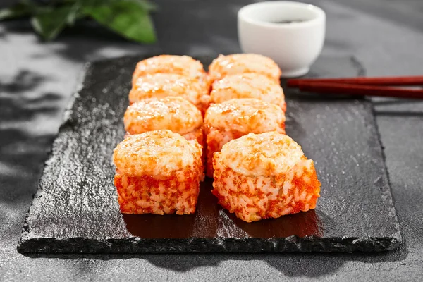 Baked Maki sushi on dark slate. Hot california maki with tobiko. Sushi roll with masago caviar, cheese topped baked oven. Style concept japanese menu with black background, leaves and hard shadow