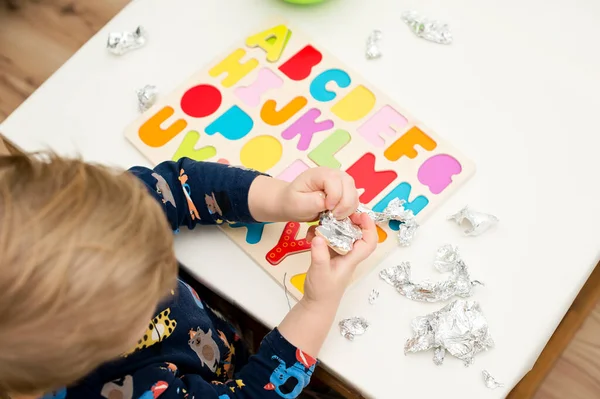 Tahta alfabe harfleriyle oynayan iki yaşında bir çocuk. Folyoya sarılı mektuplar. Entellektüel oyun, erken eğitim için anaokulu uygulaması. Sözlü ve bellek alıştırması.