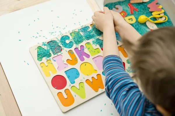 kid learning to read alphabet through game. Sensory activity with colored rice and wooden letters. Educations at home, pre-school education, Montessori methodology