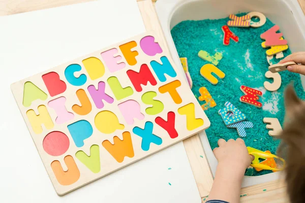 kid learning to read alphabet through game. Sensory activity with colored rice and wooden letters. Educations at home, pre-school education, Montessori methodology