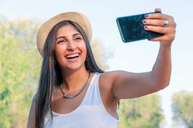 Şapkalı güzel genç bir kadın telefonuyla kendini fotoğraflıyor. Güzel gülümseyen beyaz bir genç kız güneşli bir yaz gününde dışarıda selfie çekiyor.