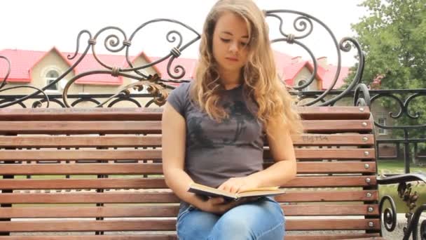 A young girl reading a book on a bench. Summer city park — Stock Video