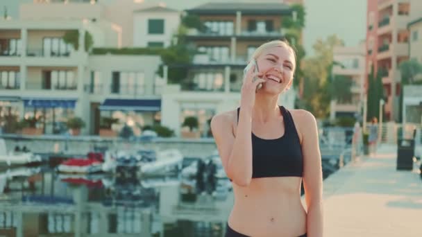 Smiling woman in yoga set speaking on smartphone in city sea port — Stock videók