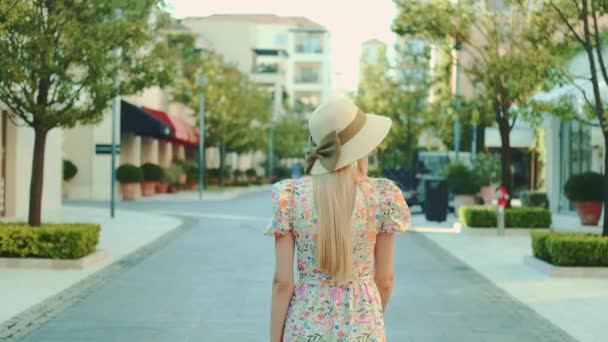 Back view of slim blonde woman in floral dress and summer hat walking — Stock videók