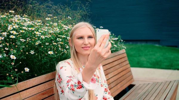 Lovely young female using smartphone to make video call while sitting on bench on living roof of huge mall. — Stock Photo, Image