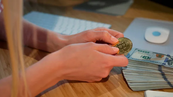 Female hands holding bitcoin. Crop view of hands of woman holding shiny metal bitcoin sitting at wooden desk with wad of cash nearby at night.