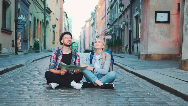 Young tourists couple using map, sitting on pavement and admiring historical surroundings