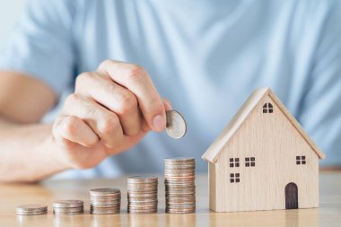 Elin bozuk para yığınına, ahşap masa üzerine ahşap ev koyuyor. Ev almak için para biriktirmek, ev kredisi konsepti.