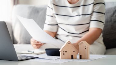 Küçük Tahta Ev Modeli ile birlikte ev kredisi ödemelerini hesap makinesi ve dizüstü bilgisayar kullanarak ev kredisi ödemelerini hesaplayan kadın, emlak, ev kredisi kavramları satın almayı planlıyor..