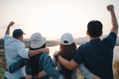 Kollarını kaldırmış bir grup insan dağın arka planında güneşin doğuşuna bakıyor. Mutluluk, başarı, dostluk ve toplum kavramları.