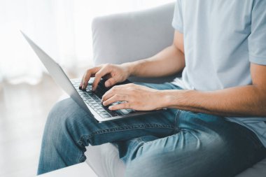 Evdeki kanepede oturan genç adam internet üzerinden çalışmak için dizüstü bilgisayarın klavyesine yazı yazıyor. İnternet, serbest yazar, rahat, modern iş hayatı sürüyor, evde çalışıyor.