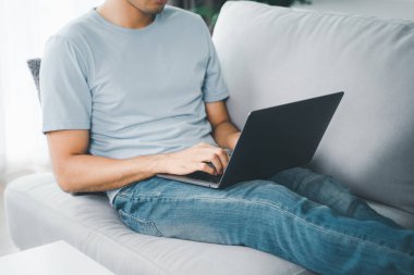 Evdeki kanepede oturan genç adam internet üzerinden çalışmak için dizüstü bilgisayarın klavyesine yazı yazıyor. İnternet, serbest yazar, rahat, modern iş hayatı sürüyor, evde çalışıyor.
