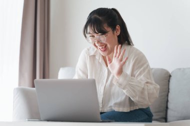 Young Asian woman using laptop computer for online video conference call waving hand making hello gesture on the couch in living room.