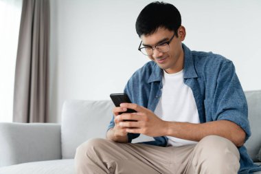 Happy Asian man using smartphone on sofa in living room at home, searching data and social media on internet.
