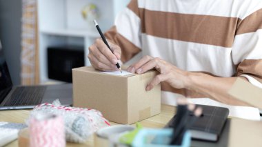 Genç Asyalı müşteri bilgilerini ve adreslerini not defterine ya da kutuya yazarak bilgilere göre nakliyeye hazırlanıyor. Paketleme kutusu, çevrimiçi satış, serbest çalışma..