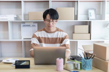 Genç adam, ürün siparişi veren müşterilerle sohbet etmek için dizüstü bilgisayar kullanıyor, evde serbest çalışma, arka planda ambalaj, çevrimiçi satış, küçük işletme sahibi, çevrimiçi alışveriş KOBİ girişimcisi.