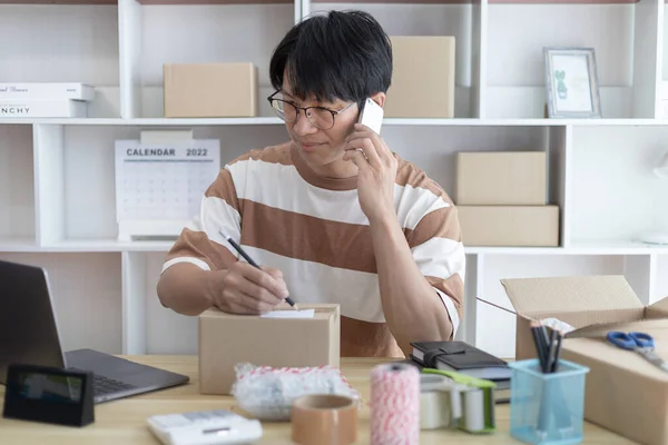 Asian man is taking online orders from a phone and chatting with customers to confirm their order, Selling products online or doing freelance work at home, Working at home and owning businesses.