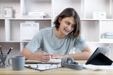 Asyalı adam not defterine not alıyor evde tablette okurken, görüntülü sohbet, online iletişim, Evde kal, Yeni Normal, Mesafeli Öğrenme