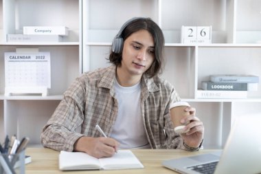 Asyalı adam evde dizüstü bilgisayarda çalışırken not alıyor, görüntülü sohbet, online iletişim, Evde kal, Yeni Normal, Mesafe Öğrenme, Sosyal Mesafe Öğrenme, Çevrimiçi Öğrenme.