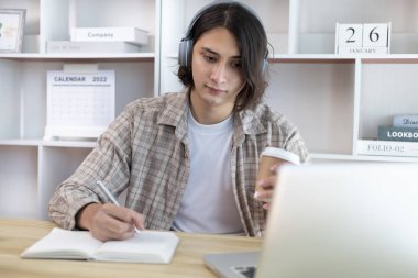 Asyalı adam evde dizüstü bilgisayarda çalışırken not alıyor, görüntülü sohbet, online iletişim, Evde kal, Yeni Normal, Mesafe Öğrenme, Sosyal Mesafe Öğrenme, Çevrimiçi Öğrenme.