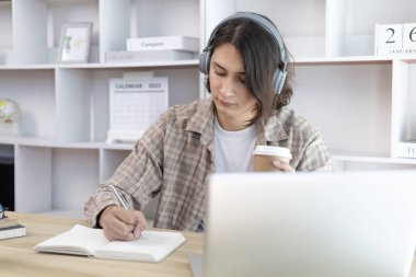 Asyalı adam evde dizüstü bilgisayarda çalışırken not alıyor, görüntülü sohbet, online iletişim, Evde kal, Yeni Normal, Mesafe Öğrenme, Sosyal Mesafe Öğrenme, Çevrimiçi Öğrenme.