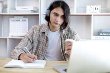Asyalı adam evde dizüstü bilgisayarda çalışırken not alıyor, görüntülü sohbet, online iletişim, Evde kal, Yeni Normal, Mesafe Öğrenme, Sosyal Mesafe Öğrenme, Çevrimiçi Öğrenme.