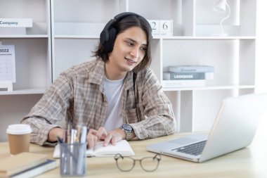 Asyalı adam evde dizüstü bilgisayarda çalışırken not alıyor, görüntülü sohbet, online iletişim, Evde kal, Yeni Normal, Mesafe Öğrenme, Sosyal Mesafe Öğrenme, Çevrimiçi Öğrenme.