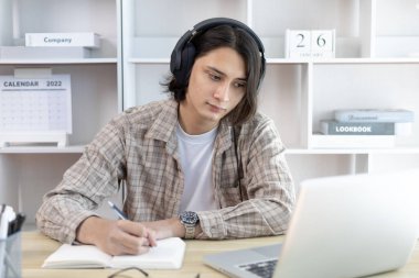 Asyalı adam evde dizüstü bilgisayarda çalışırken not alıyor, görüntülü sohbet, online iletişim, Evde kal, Yeni Normal, Mesafe Öğrenme, Sosyal Mesafe Öğrenme, Çevrimiçi Öğrenme.