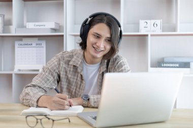 Asyalı adam evde dizüstü bilgisayarda çalışırken not alıyor, görüntülü sohbet, online iletişim, Evde kal, Yeni Normal, Mesafe Öğrenme, Sosyal Mesafe Öğrenme, Çevrimiçi Öğrenme.