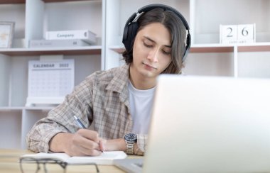 Asyalı adam evde dizüstü bilgisayarda çalışırken not alıyor, görüntülü sohbet, online iletişim, Evde kal, Yeni Normal, Mesafe Öğrenme, Sosyal Mesafe Öğrenme, Çevrimiçi Öğrenme.