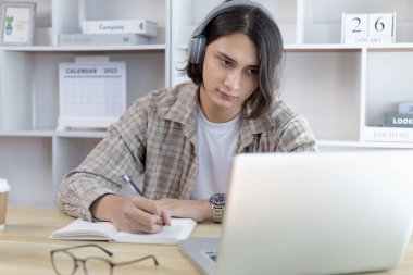 Asyalı adam evde dizüstü bilgisayarda çalışırken not alıyor, görüntülü sohbet, online iletişim, Evde kal, Yeni Normal, Mesafe Öğrenme, Sosyal Mesafe Öğrenme, Çevrimiçi Öğrenme.