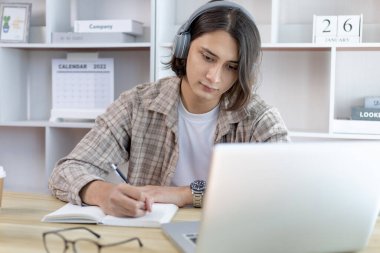 Asyalı adam evde dizüstü bilgisayarda çalışırken not alıyor, görüntülü sohbet, online iletişim, Evde kal, Yeni Normal, Mesafe Öğrenme, Sosyal Mesafe Öğrenme, Çevrimiçi Öğrenme.