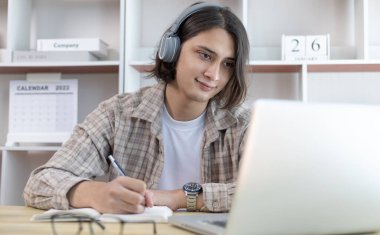 Asyalı adam evde dizüstü bilgisayarda çalışırken not alıyor, görüntülü sohbet, online iletişim, Evde kal, Yeni Normal, Mesafe Öğrenme, Sosyal Mesafe Öğrenme, Çevrimiçi Öğrenme.