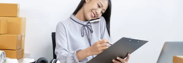 Asian businesswoman is taking online orders from a phone and chatting with customers to confirm their order, Selling products online or doing freelance work at home concept.