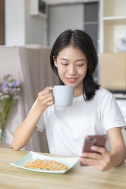 Asyalı kadın kahvaltısını kraker (sağlıklı tam tahıl) ve meyve suyu ile yer, telefonunuzu ya da cep telefonunuzu sabah haberlerini aramak için kullanın, apartman zemininde küçük bir oda, uyandırma aktiviteleri.