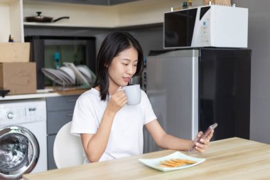 Asyalı kadın kahvaltısını kraker (sağlıklı tam tahıl) ve meyve suyu ile yer, telefonunuzu ya da cep telefonunuzu sabah haberlerini aramak için kullanın, apartman zemininde küçük bir oda, uyandırma aktiviteleri.