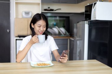Asyalı kadın kahvaltısını kraker (sağlıklı tam tahıl) ve meyve suyu ile yer, telefonunuzu ya da cep telefonunuzu sabah haberlerini aramak için kullanın, apartman zemininde küçük bir oda, uyandırma aktiviteleri.