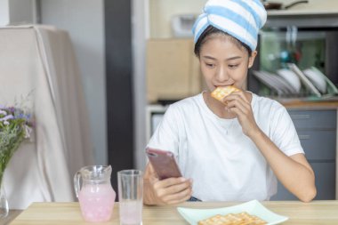 Asyalı kadın kahvaltısını kraker (sağlıklı tam tahıl) ve meyve suyu ile yer, telefonunuzu ya da cep telefonunuzu sabah haberlerini aramak için kullanın, apartman zemininde küçük bir oda, uyandırma aktiviteleri.