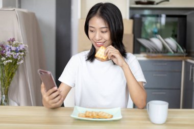 Asyalı kadın kahvaltısını kraker (sağlıklı tam tahıl) ve meyve suyu ile yer, telefonunuzu ya da cep telefonunuzu sabah haberlerini aramak için kullanın, apartman zemininde küçük bir oda, uyandırma aktiviteleri.