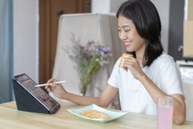 Asyalı kadın kahvaltısını kraker (sağlıklı tam tahıl) ve meyve suyu ile yer, telefonunuzu ya da tabletinizi sabah haberlerini aramak için kullanın, apartman zemininde küçük bir oda, uyandırma aktiviteleri