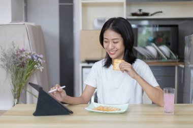 Asyalı kadın kahvaltısını kraker (sağlıklı tam tahıl) ve meyve suyu ile yer, telefonunuzu ya da tabletinizi sabah haberlerini aramak için kullanın, apartman zemininde küçük bir oda, uyandırma aktiviteleri