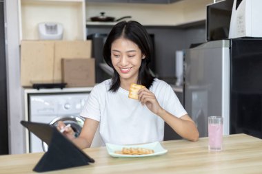 Asyalı kadın kahvaltısını kraker (sağlıklı tam tahıl) ve meyve suyu ile yer, telefonunuzu ya da tabletinizi sabah haberlerini aramak için kullanın, apartman zemininde küçük bir oda, uyandırma aktiviteleri