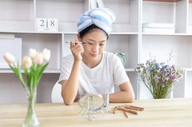 Asyalı bir kadın işe gitmek için saçını havluya sarıp makyaj yapıyor, makyaj yapıyor, sabah duş aldıktan sonra güzellik, yüz bakımı ve makyaj malzemesi, makyaj aynası..