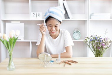 Asyalı bir kadın işe gitmek için saçını havluya sarıp makyaj yapıyor, makyaj yapıyor, sabah duş aldıktan sonra güzellik, yüz bakımı ve makyaj malzemesi, makyaj aynası..