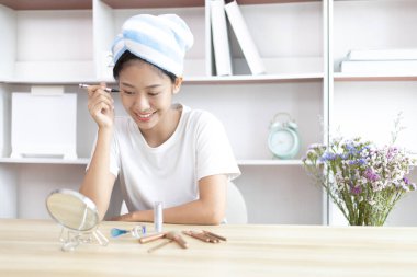 Asyalı bir kadın işe gitmek için saçını havluya sarıp makyaj yapıyor, makyaj yapıyor, sabah duş aldıktan sonra güzellik, yüz bakımı ve makyaj malzemesi, makyaj aynası..