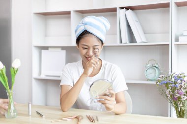 Asyalı bir kadın işe gitmek için saçını havluya sarıp makyaj yapıyor, makyaj yapıyor, sabah duş aldıktan sonra güzellik, yüz bakımı ve makyaj malzemesi, makyaj aynası..
