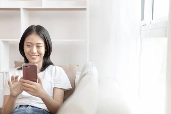 Asian woman sitting with smart phone on sofa in living room, Looking at tablet screen, Relaxing at home, Working through the Internet communication system, Happy working lifestyle.