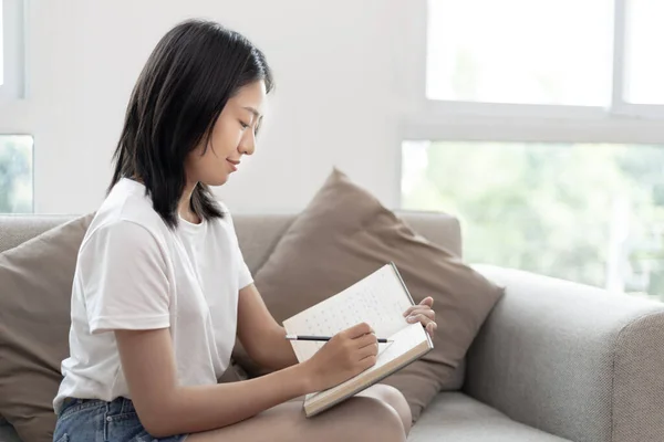 Asian woman writes notebook or a diary on her bedroom couch at home during the holidays, New work lifestyle, Relaxation, Happy time, Living at home, Sit comfortably on vacation, Comfortable corner.