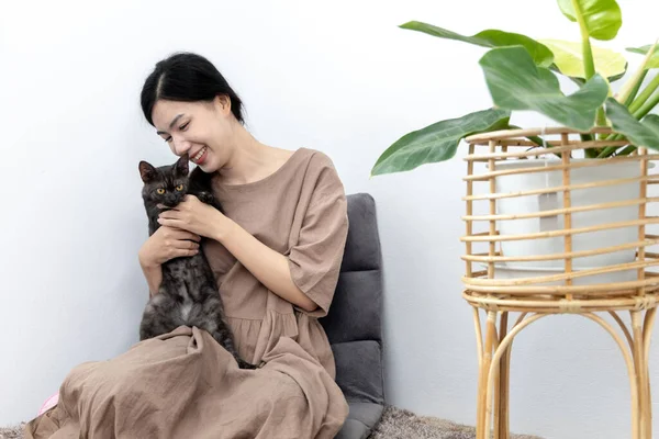 Asyalı kadın mutluca kedisiyle oturma odasında oynuyor, sevgili evcil hayvan, en iyi arkadaş, mutlu bölge, evde tatiller, rahatla ve eğlen, kediyle halının üzerinde oturuyor,.
