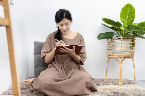 Asian woman sits in her living room writing a diary or notebook on the carpet, Weekends or holidays at home, Relaxation, Happy time, Living at home, Sit comfortably on vacation, Comfortable corner.